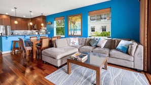 A modern living room with a blue accent wall, a wooden floor, a beige sofa, a glass coffee table, and a kitchen area with wooden cabinets and pendant lights.