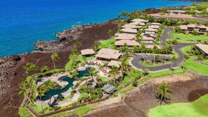 An aerial view of a resort with multiple bungalow-style buildings, a central pool area, and a view of the ocean.