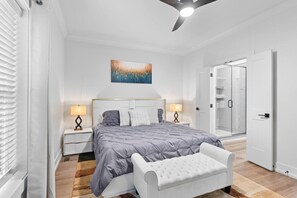 Un dormitorio con cama, mesitas de noche, un banco, un ventilador de techo y un vidrio que da a un baño.