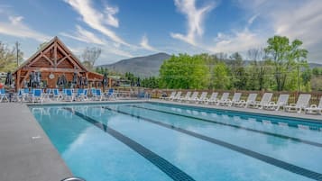 Une piscine avec des chaises longues et des parasols, un chalet en bois et un paysage montagneux en arrière-plan.