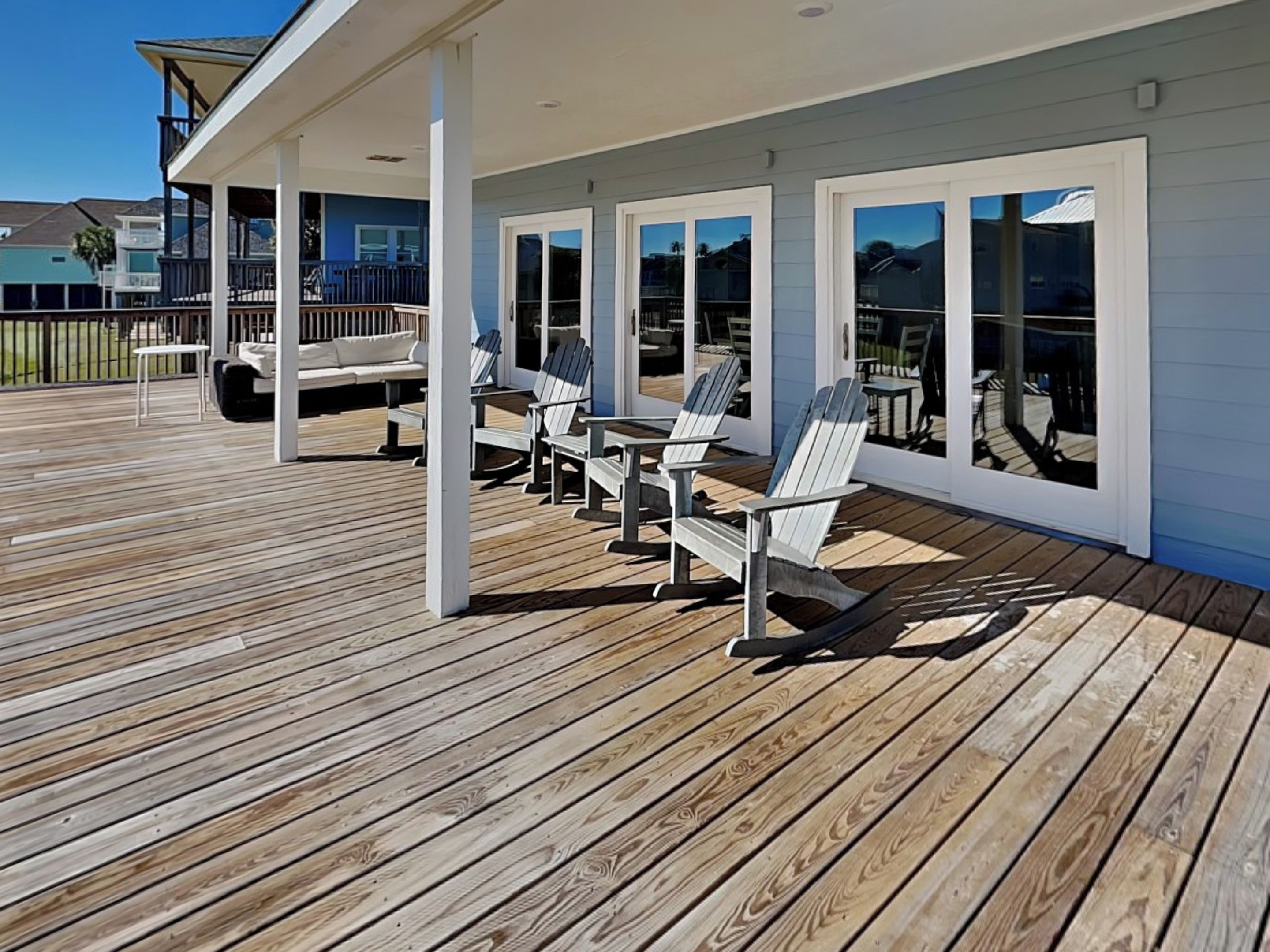 A covered outdoor deck with wooden flooring, white Adirondack chairs, and large windows.