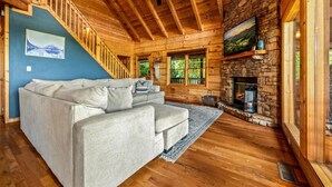 Interior acogedor de una cabaña con escalera de madera, chimenea de piedra y un ventanal con vistas a la vegetación.