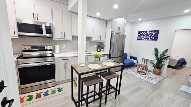 A modern kitchen with stainless steel appliances, a dining area with a wooden table and stools, and a living area with a sofa and a decorative plant.