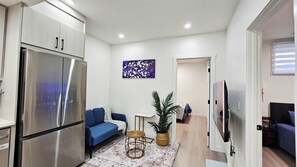 Espaço compacto para viver com cozinha compacta, sofá azul, mesa pequena, planta em vaso e TV de tela plana na parede.