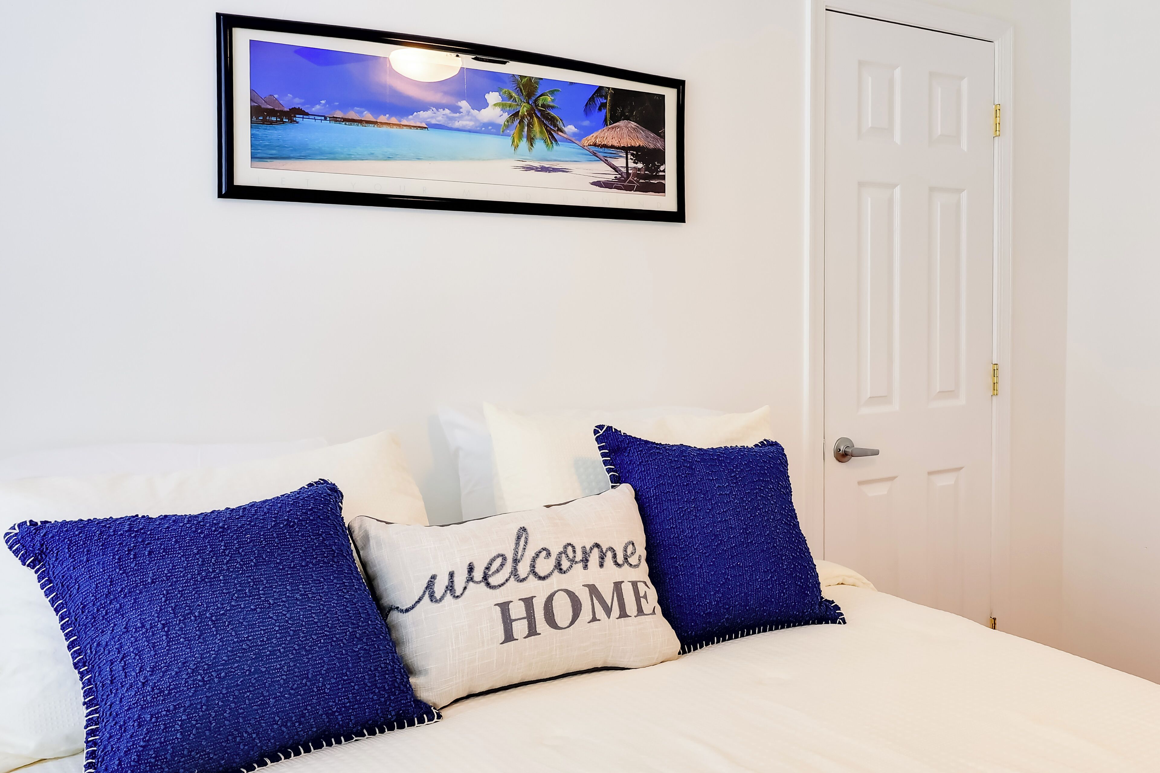 A bedroom with a bed, a framed picture of a beach scene, and a door.