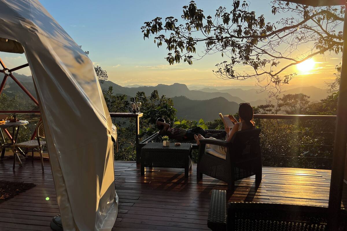 Ein mit Zelten überspanntes Areal mit Blick auf Berge und einen Sonnenuntergang. Wicker Möbel und ein Tisch mit Flasche und Gläsern sind ebenfalls vorhanden.