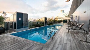 A rooftop pool with a clear view of the city skyline, a modern building with a glass facade, and a lounge area with sun loungers. - Blissful Sanctuary 1BR Balcony w/ Canal View in vibrant Downtown Dubai (Dubai)