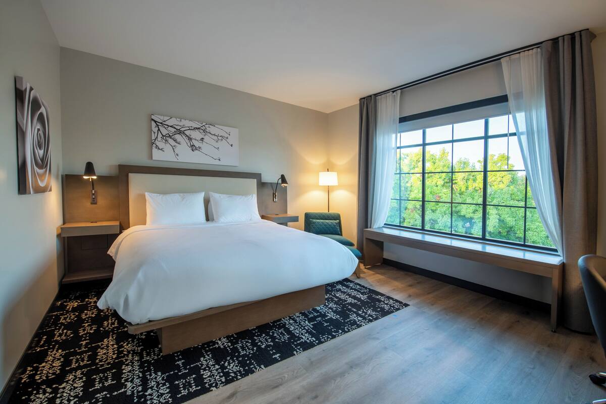 A modern hotel room with a large bed, two bedside tables, a desk, and a window with curtains.