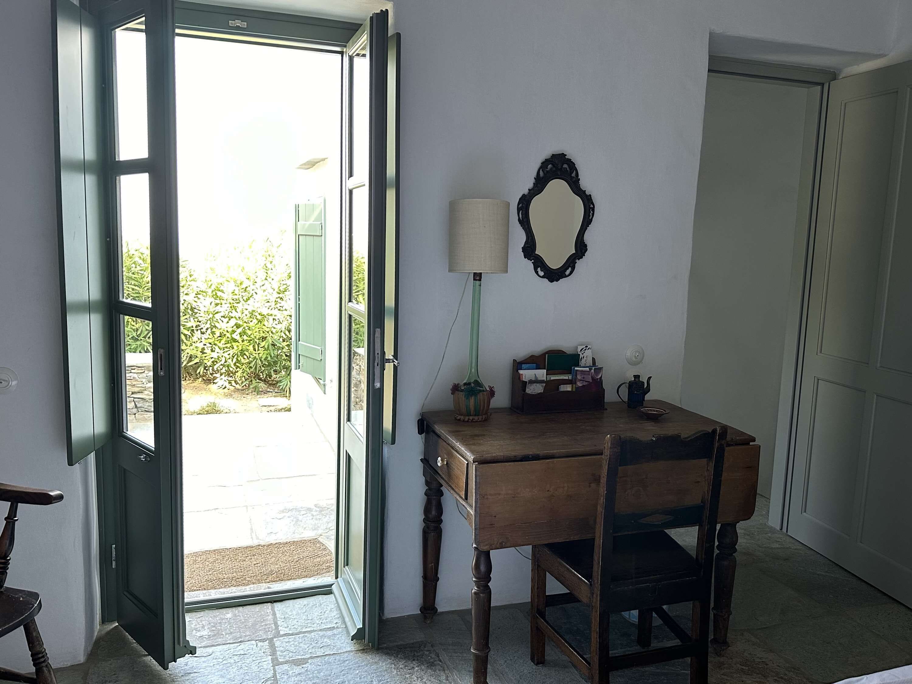 A room with a wooden table, a chair, a lamp, a mirror, and a door leading outside.