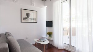A modern living room with a grey sofa, a small white table with a magazine and remote, a framed picture on the wall, and a large window with sheer curtains.