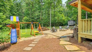 Ein Spielplatz mit Rutsche und Schaukeln, umgeben von Wald und einer Holzterrasse mit Sitzgelegenheiten.