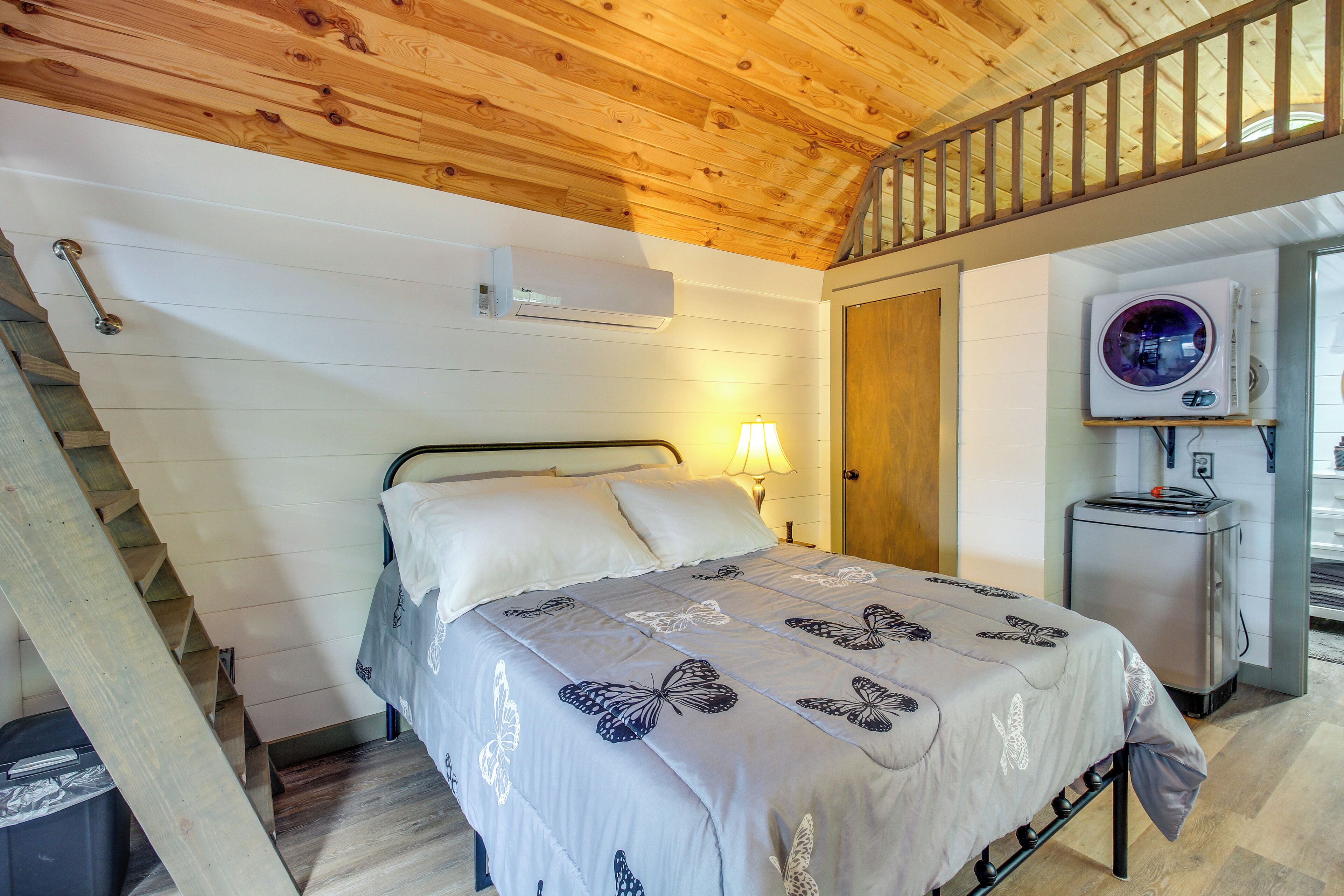 A bedroom with a bed, a nightstand, a lamp, an air conditioning unit, and a small refrigerator.