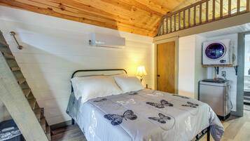 A bedroom with a bed, a nightstand, a lamp, an air conditioning unit, and a small refrigerator.