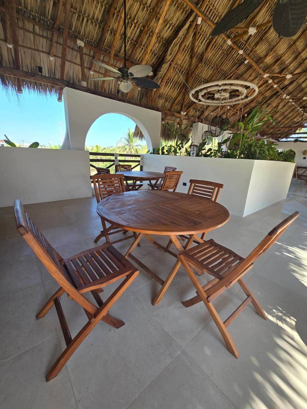 Een buitenterras met een ronde houten tafel en stoelen, een rieten dak en uitzicht op de omgeving.