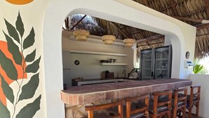 Un espace bar avec un comptoir en pierre et des tabourets en bois, une cuisine visible à travers une arche, et un toit de chaume au-dessus.