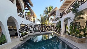 Una piscina con muelle de madera y un puente, rodeada de edificios blancos con techos de paja y palmeras.