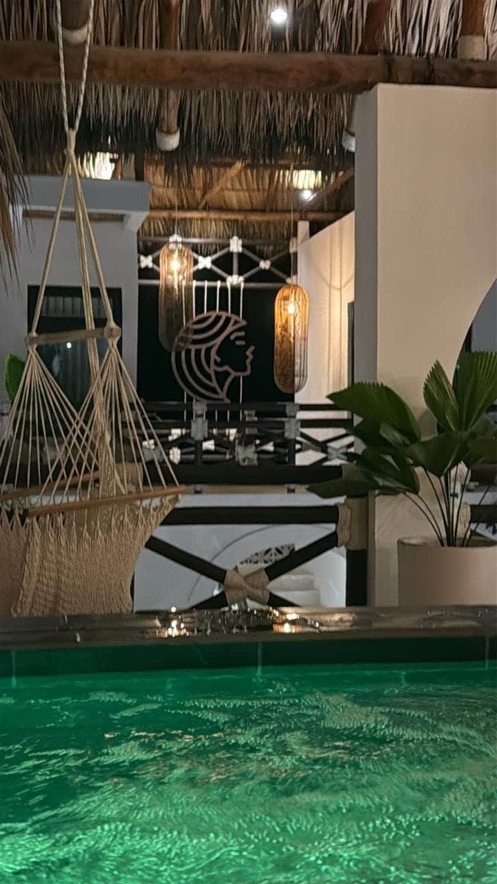 Un área de piscina con techo de paja, lámparas colgantes de macramé y una pared decorativa con cabeza de león.