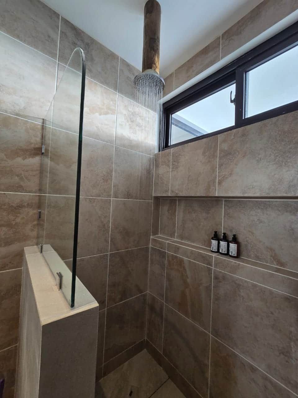 Eine Dusche mit Glasabtrennung, einer senkrechten Brause und einem Fenster darüber.