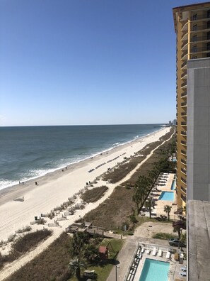 Blick auf den Strand von einem Hochhaus, mit einem langen Sandstrand, einer Holzpromenade und einem Poolbereich.
