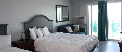 Quarto de hotel com uma cama grande, uma cama menor, uma mesa de cabeceira, cômoda, espelho, aparelho de ar condicionado e vista para o exterior.