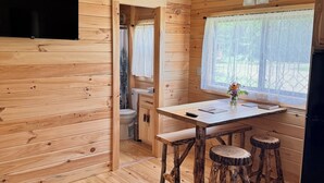 L’intérieur d’une cabane en bois comprend une table à manger, des tabourets, un coin cuisine et une fenêtre avec des rideaux.