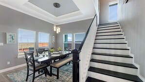 Un espace repas comprenant une longue table, des chaises et un banc. On y trouve un escalier avec des marches noires et une rampe blanche.