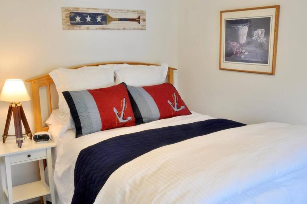 Una cama bien tendida con una colcha azul marino y blanca a rayas, un cabecero de madera y dos almohadas con motivos de ancla. Se ve una mesita de noche con una l ámpara y un reloj.