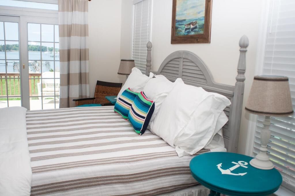 Un dormitorio con cama, ropa de cama a rayas, almohada colorida, mesita de noche, lámpara y vista a un lago a través de una ventana.