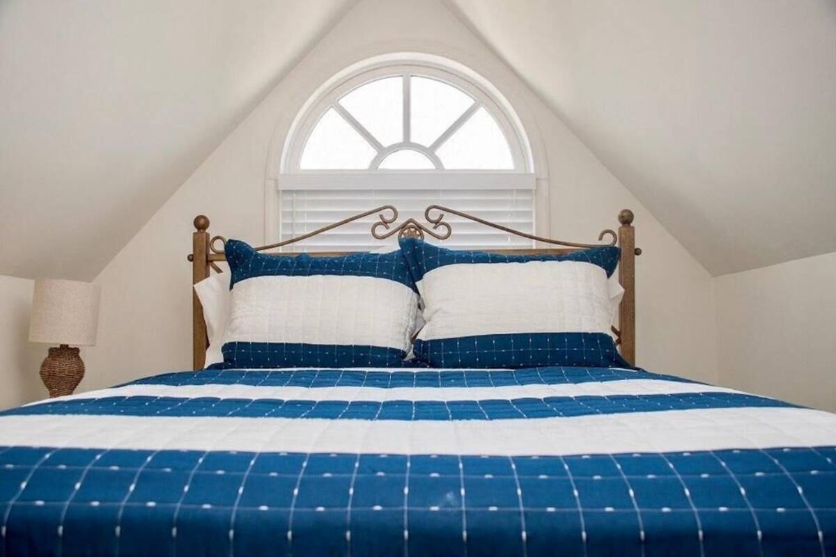 Un dormitorio con una cama en la esquina, una ventana con contraventanas blancas y una lámpara en la mesita de noche.