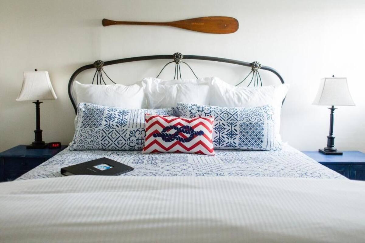 Una cama bien tendida con almohadas estampadas y una almohada decorativa, un remo de madera en la cabecera y lámparas de noche a ambos lados.