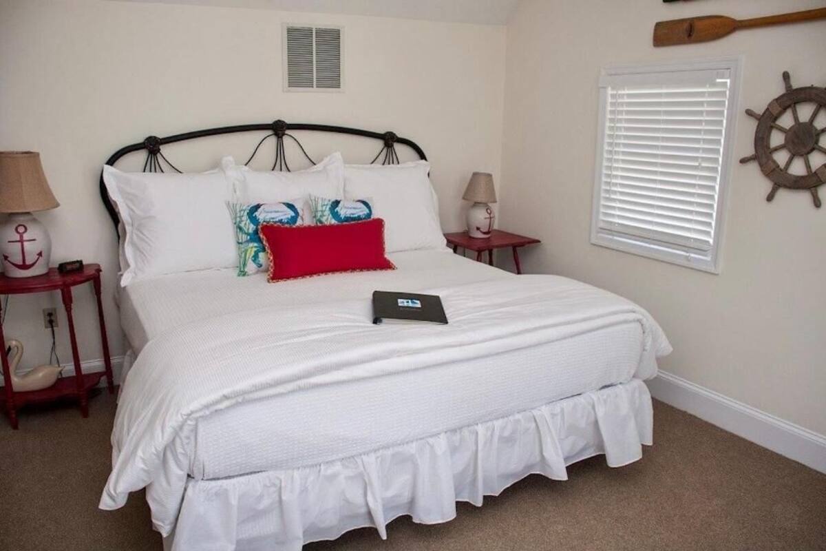 Una cama cuidadosamente arreglada con ropa de cama blanca y un cojín rojo, un marco de cama de madera, dos mesitas de noche con lámparas, una ventana con persianas y un timón de barco montado en la pared.