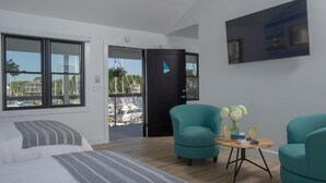 A modern hotel room with a large bed, two teal armchairs, a small round table with drinks, and a large window offering a view of the marina. - Inn at Haven Harbour (Rock Hall)