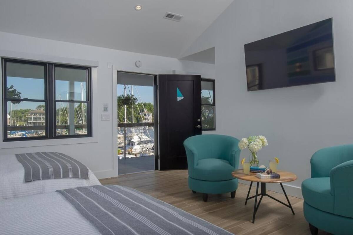 Habitación de hotel moderna con una cama grande, dos sillones color turquesa, una mesita redonda con bebidas y un ventanal con vistas al puerto deportivo.