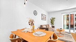 Un coin repas avec une table en bois dressée pour deux personnes, un vase contenant des fleurs séchées et une bouteille de vin. On aperçoit un canapé et une plante au fond.