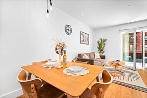 A dining area with a wooden table set for two, a vase with dried flowers, and a bottle of wine. There is a sofa and a plant in the background. - Luxury Apartment - Balcony - Fast WiFi - Netflix (Birmingham)