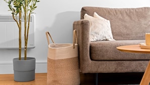 Ein Wohnzimmer mit grauem Sofa, einem runden Holztisch, einem grauen gewebten Korb und einer Topfpflanze.