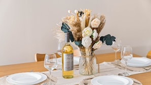 Ein Esstisch, gedeckt mit weißen Tellern, einer Flasche Pinot Grigio und einer Vase mit Blumen und Pampasgras.