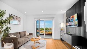 Ein modernes Wohnzimmer mit grauem Sofa, einem Couchtisch, einem Fernseher und Blick auf die Stadt durch große Glaswände.