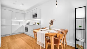 Une cuisine moderne avec des meubles blancs, une table à manger en bois pour quatre personnes et une étagère noire agrémentée de plantes.