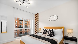 Une chambre moderne avec un grand lit, une vue sur un immeuble en briques, une œuvre d’art murale originale et une lampe contemporaine.