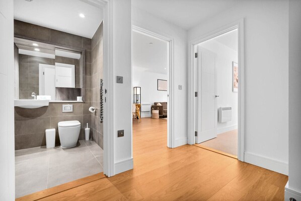 Un baño moderno con un espejo grande, un inodoro y un lavabo. El baño tiene piso de baldosas y piso de madera que conduce a un área de estar con un sofá y una mesa de centro.