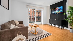 Un salon moderne avec un canapé, une petite table ronde, un téléviseur fixé au mur et une grande fenêtre donnant sur un immeuble en briques.