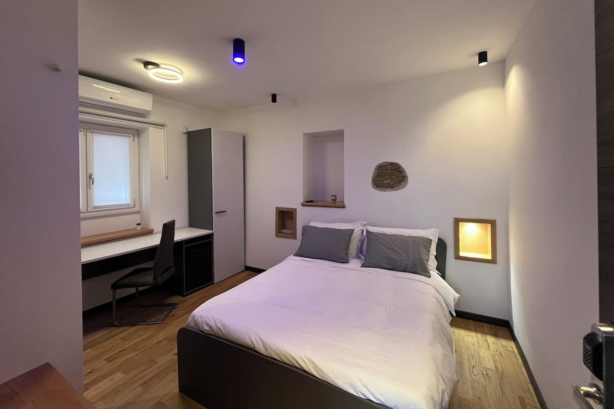 A bedroom with a bed, a desk, and a chair. There is a window with blinds, a wall-mounted shelf, and a small stone decoration.