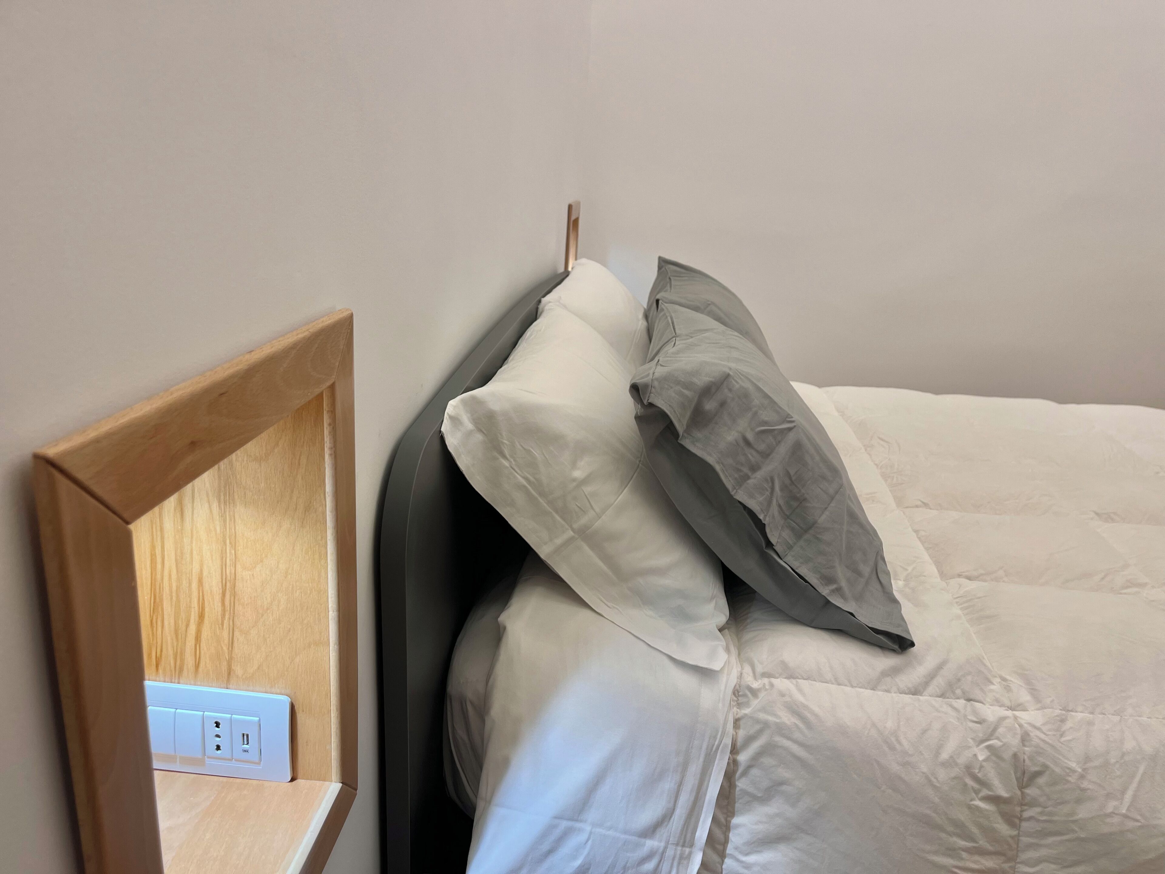 Een net opgemaakt bed met witte en grijze kussens, een houten hoofdbord en een stopcontact met een stekkerdoos.
