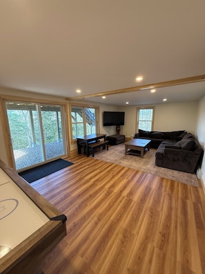 A spacious living room with a wooden floor, a large flat-screen TV, a sofa, a coffee table, and a dining table with chairs.