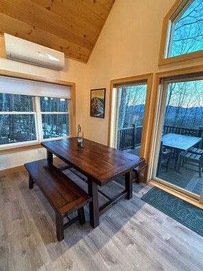A wooden interior with a dining table, benches, a mounted air conditioner, and large windows offering views of a snowy landscape. - Gorgeous Mountainside Chalet in Jackson, NH (Jackson)