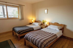 A room with two wooden beds, each with a headboard, a nightstand, and a lamp. There is a window with curtains, a small wall-mounted picture, and a wooden floor.