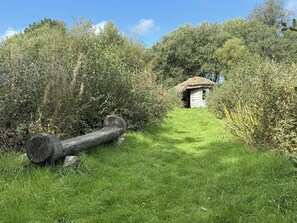 Ein rustikaler Holzbau mit einem strohgedeckten Dach, umgeben von dichter Vegetation und einem grasbewachsen Pfad.