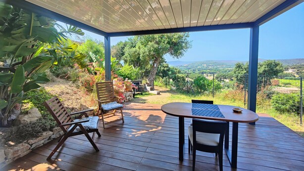 A covered outdoor patio with wooden flooring, a table with chairs, and a view of a lush landscape with trees and hills. - Mimosa house, T2 48 M2, quiet location with unobstructed view, 9min from beaches (Bormes-les-Mimosas)