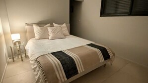 A neatly made bed with a striped blanket, a bedside table with a lamp, and a window with blinds. - Mimosa house, T2 48 M2, quiet location with unobstructed view, 9min from beaches (Bormes-les-Mimosas)